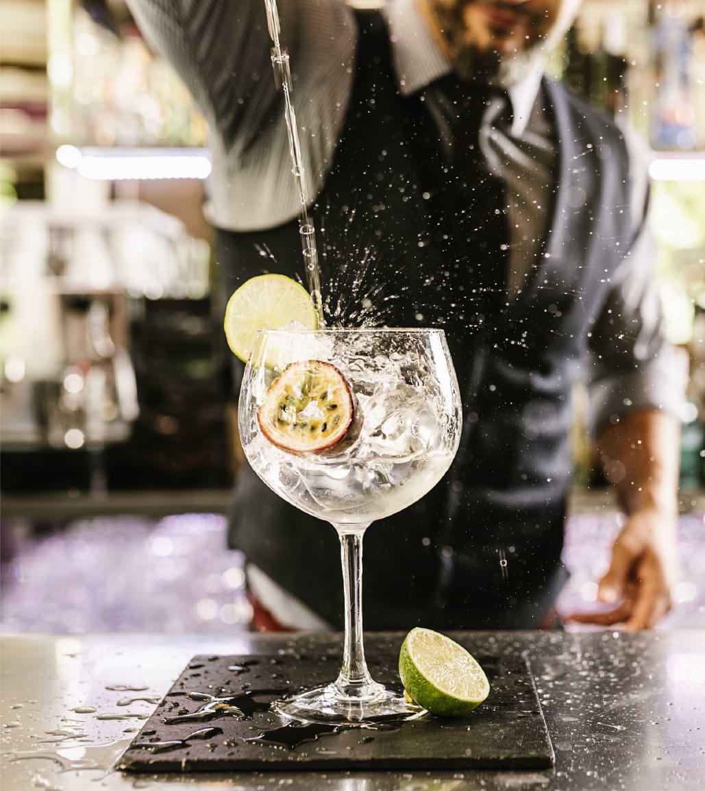 A bartender, out of focus, pouring a liquid into a glass with ice and a lime from out of frame.
